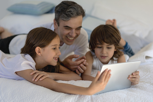Excited kids playing with daddy using a tablet while lying down on bed at home