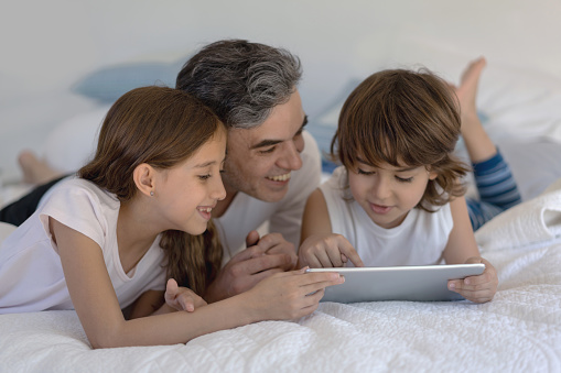 Happy single daddy with his two kids lying down on bed all playing with a tablet smiling