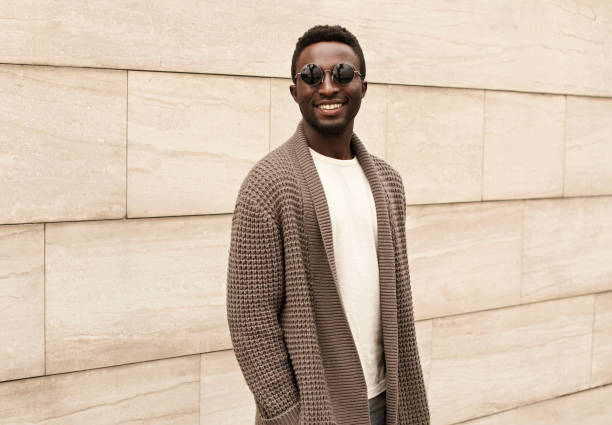 stilvolle lächelnde afrikanische mann trägt braun gestrickte strickjacke und sonnenbrille auf der stadtstraße über ziegelwand hintergrund - cardigan stock-fotos und bilder