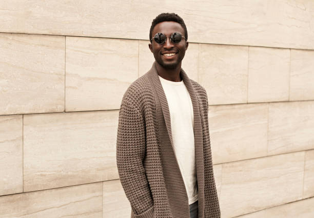 elegante hombre africano sonriente con cardigan de punto marrón y gafas de sol en la calle de la ciudad sobre fondo de pared de ladrillo - cardigan men african ethnicity african descent fotografías e imágenes de stock