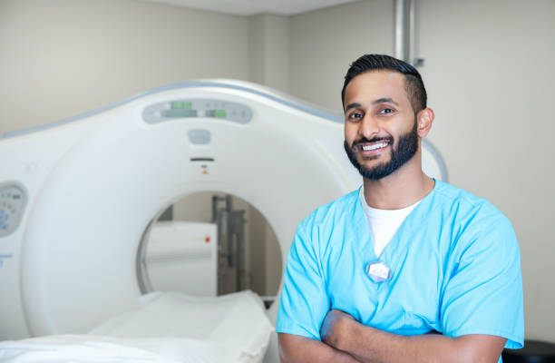 Young adult radiologist smiles while standing next to CT machine Young adult radiologist smiles while standing next to CT machine cat scan stock pictures, royalty-free photos & images