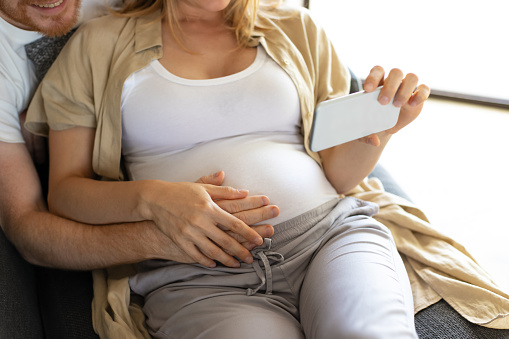 Expectant parents touching belly and using smartphone together. Pregnant young woman spending leisure time at home. Digital communication concept