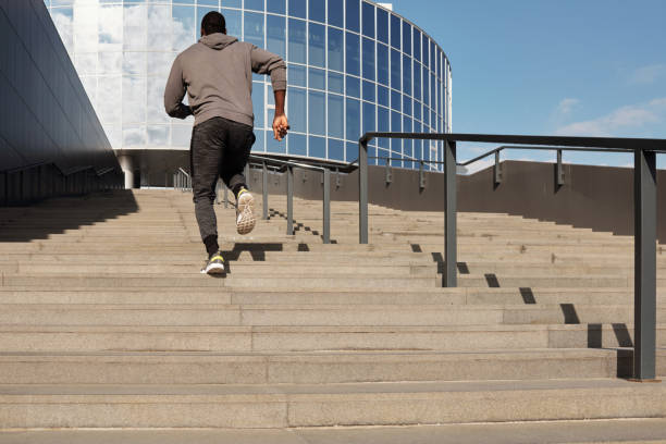 athlète noir méconnaissable dans les vêtements de sport courant vers le haut des escaliers vers le centre d'affaires avec la façade en verre, vue arrière d'en bas - exercising motivation looking up african descent photos et images de collection