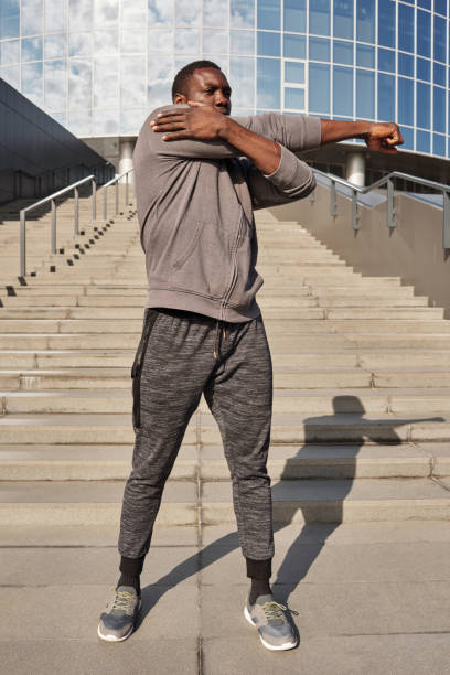 istock Young African American sportsman in activewear doing warming up exercises while standing outdoors against modern business center 1198379850