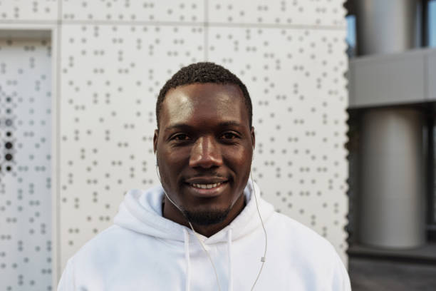 retrato de cintura arriba del joven guapo hombre negro en sudadera con capucha blanca con auriculares sonriendo a la cámara mientras se para contra el centro de negocios moderno - waistup fotografías e imágenes de stock