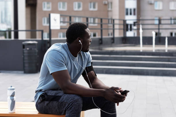 Young athletic African American man in sportswear resting on bench in city center and listening to music through earphones after workout Young athletic African American man in sportswear resting on bench in city center and listening to music through earphones after workout center athlete stock pictures, royalty-free photos & images