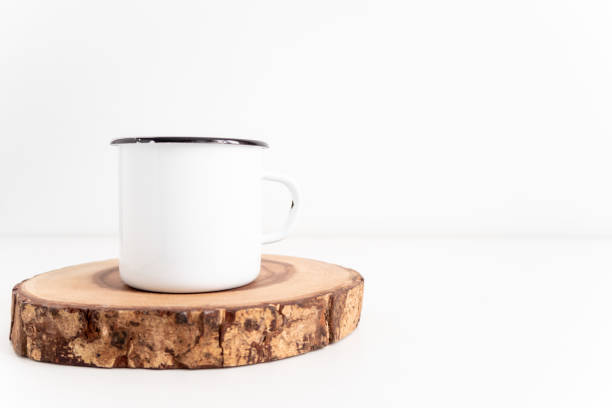 Enamel mug mock up with wooden cut tree section on white table. Boho design of tin cup on white background with copy space for image or text stock photo