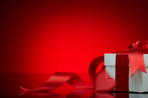 Gift box and red satin heart on a red background. Valentine's Day card with copy space. Design element for romantic greeting card, wedding invitation, Women's Day postcard
