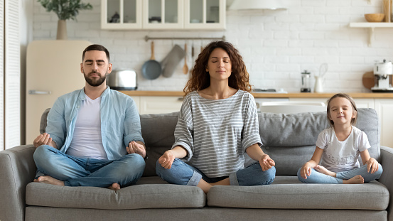 Horizontal banner serene couple and little daughter sitting on sofa in lotus position closed eyes do meditation breathing technique in living room, keep calm, healthy life habits and lifestyle concept