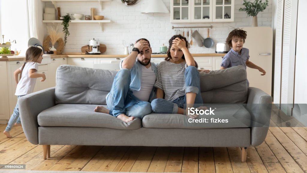 Parents tired of noisy children who are running and shouting Tired mother and father sitting on couch feels annoyed exhausted while noisy little daughter and son shouting run around sofa where parents resting. Too active hyperactive kids, need repose concept Family Stock Photo