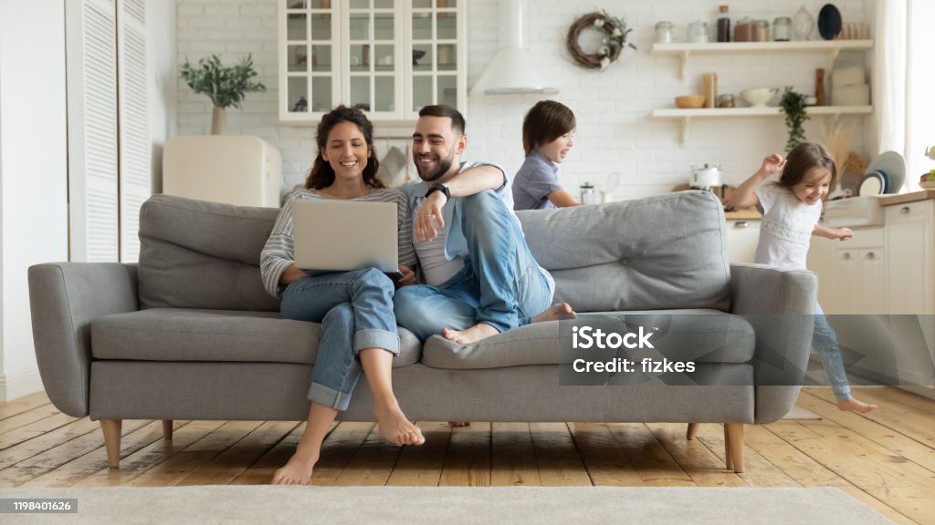 Mientras que los niños activos que dirigen a los padres descansan en el sofá usando computadora portátil - Foto de stock de Familia libre de derechos