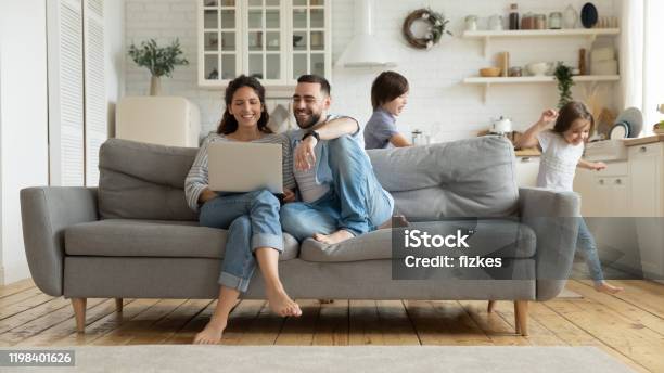 Terwijl Actieve Kinderen Running Ouders Rusten Op De Bank Met Behulp Van Laptop Stockfoto en meer beelden van Familie