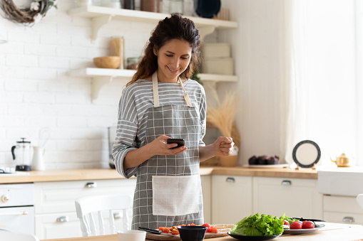 Woman wearing apron standing on domestic kitchen table full of fresh vegetables, housewife holds phone using cooking apps websites search recipes, chatting with friend distracted from food preparation