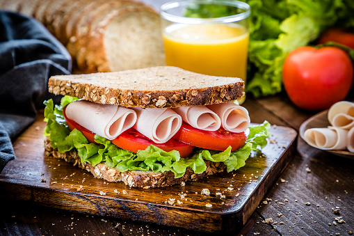 Front view of a sandwich made with ham, tomatoes, lettuce and wholemeal bread. Focus on foreground. At the background is a drinking glass full of orange juice and the ingredients for preparing the sandwich like lettuce, tomatoes, ham and bread. The sandwich is on top of a rustic wooden cutting board. Low key DSLR photo taken with Canon EOS 6D Mark II and Canon EF 24-105 mm f/4L