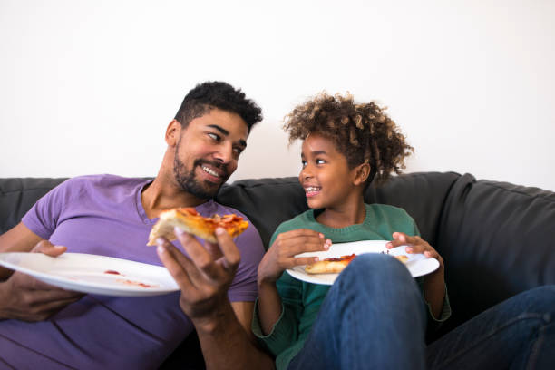 padre e hija disfrutando de la pizza en cómodo sofá de la sala de estar. - peinado desarreglado fotografías e imágenes de stock