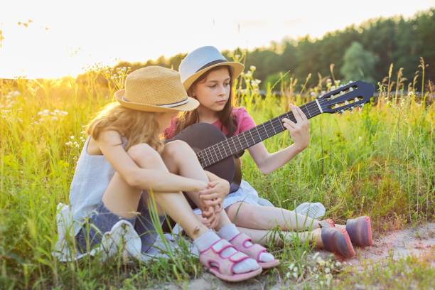 Children sitting in nature with classical guitar, two girls learning playing the guitar and singing, enjoying music and summer day