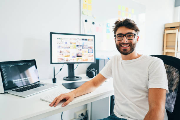 istock Young photographer looking at camera and smiling while sitting in office with laptop editing pictures 1198428309