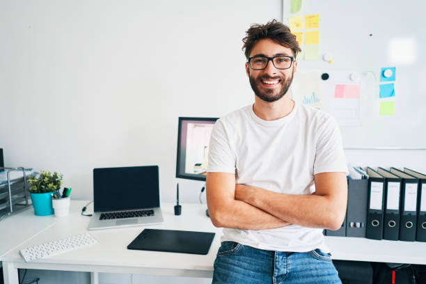 Portrait of confident graphic designer leaning on desk in office with arms crossed Portrait of confident graphic designer leaning on desk in office with arms crossed young graphic designer stock pictures, royalty-free photos & images