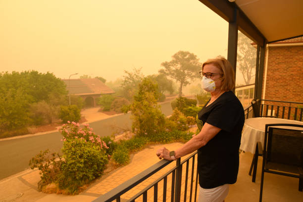 A woman, on a balcony, wears a face mask to protect herself against very dangerous level of air pollution.  A thick yellow fog coming from the nearby forest fires covers Canberra city. Canberra, Australia, 5/01/2020, Smoke coming from nearby forest fires creates high pollution and covers Canberra with a thick fog. People need to wear mask to breathe. smog stock pictures, royalty-free photos & images