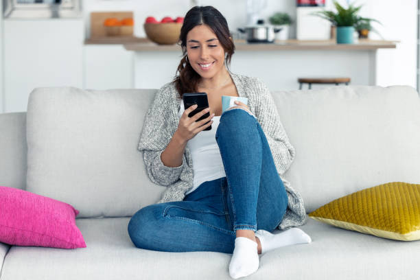 istock Pretty young woman using her mobile phone while drinking coffee on sofa at home. 1198429615