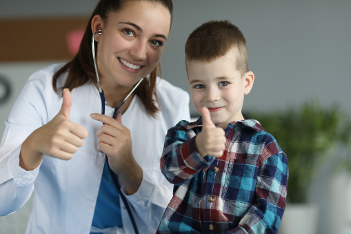 Portrait of smart pediatrician with little patient in clinic office showing thumbs up to camera. Joyful kid smiling and performing approving gesture. Medicine and healthcare concept