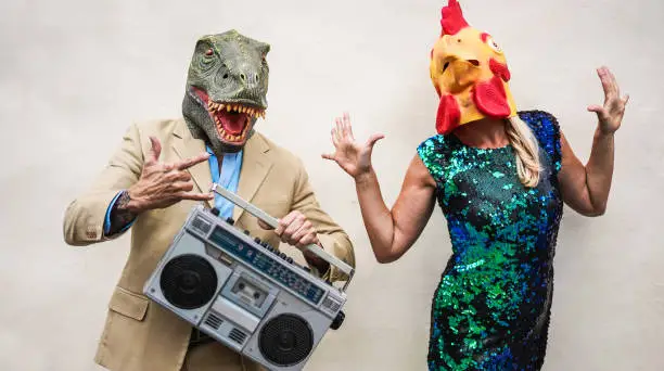 Photo of Crazy senior couple dancing at carnival party wearing t-rex and chicken mask - Old trendy people having fun listening music with boombox stereo - Absurd and funny trend concept - Focus on faces