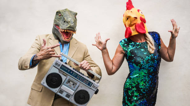 dança sênior louca dos pares no partido do carnaval que desgasta o t-rex e a máscara de galinha - povos na moda idosos que têm a música de escuta do divertimento com estéreo do boombox - conceito absurdo e engraçado da tendência - foco em caras - senior couple audio - fotografias e filmes do acervo