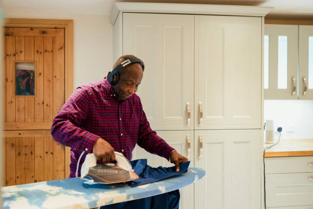 He Loves Helping Round the House A senior man ironing his clothes on an ironing board while listening to music through wireless headphones. role reversal stock pictures, royalty-free photos & images