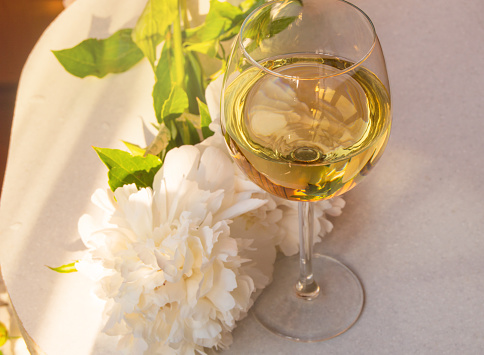 One glass of white wine and a white peony flower on a white table, top view. Relax with a glass of wine on a warm summer evening.