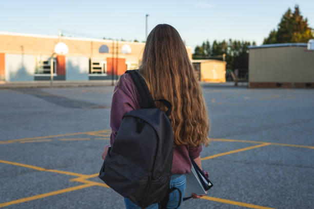 Profile of a teen girl depressed/sad at sunset in a parking lot while wearing a backpack and holding binders. stock photo