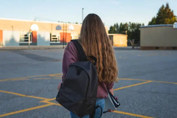 Photo of Profile of a teen girl depressed/sad at sunset in a parking lot while wearing a backpack and holding binders.
