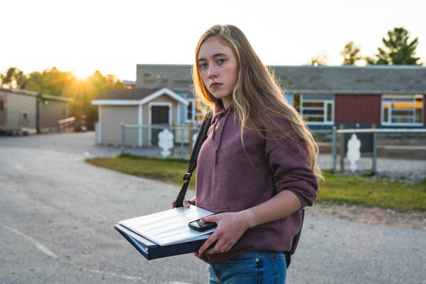 Sad/depressed teen girl/student standing in front of a school during sunset while wearing a backpack and holding binders/smartphone. stock photo