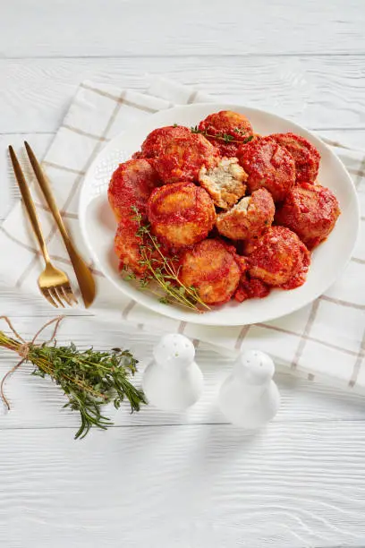 Boulettes de Poisson, Fried Fish Balls in Tomato Sauce on a white plate on a wooden table with golden fork and knife and bouquet of aromatic herbs,vertical view from above