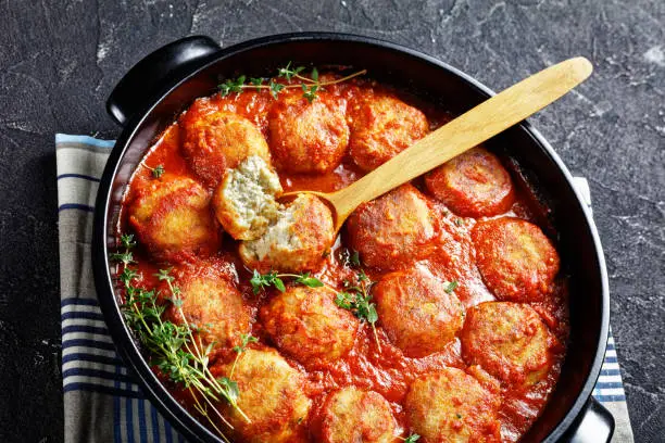 close-up of Boulettes de Poisson, Fried Fish Balls in Tomato Sauce in a black dish on a concrete table, horizontal view from above