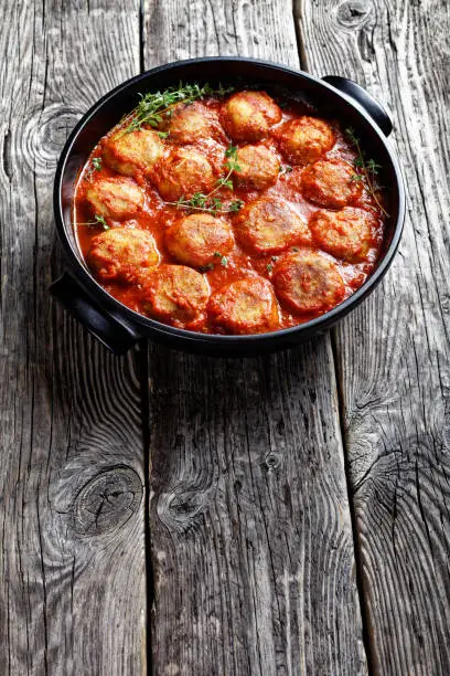 Boulettes de Poisson, Fried Fish Balls in Tomato Sauce in a black dish on a rustic wooden table, vertical view from above