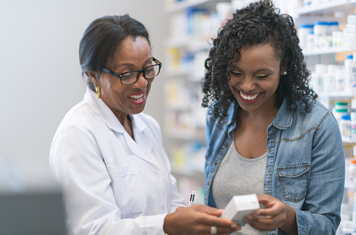 A young African woman consults a female pharmacist about an over the counter medication, while shopping in a pharmacy.  She is dressed casually in a white t-shirt and a denim jacket, while the African pharmacist is wearing a white lab coat and has her hair pulled back.