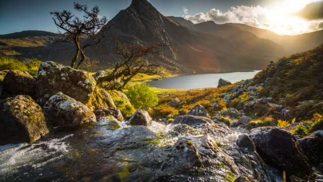 Crane shot of landscape at Snowdonia National Park in Wales. Mountain stream with fresh water in the foreground.
Tryfan mountain and Llyn Ogwen in the background. Warming evening sunlight producing sunbeams in the distance. Northern Wals, UK