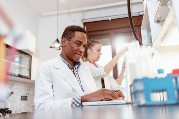 He only uses smart tools in his lab Photo of one male scientist typing on laptop with his female colleague on background. biochemist stock pictures, royalty-free photos & images