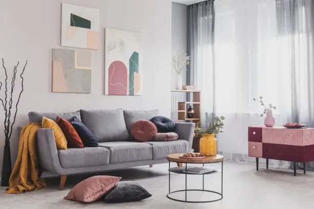 Photo of Real photo of abstract paintings hanging on white wall above a gray sofa in a living room interior with big windows