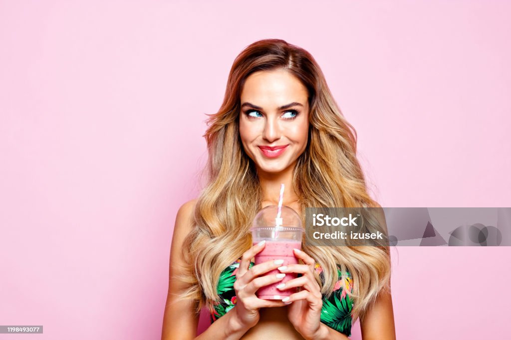 Summer portrait of young blonde woman holding smoothie Glamour portrait of beautiful long hair young woman wearing floral swimsuit, holding smoothie, looking away. Studio shot on pink background. Women Stock Photo