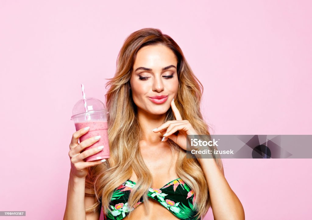 Summer portrait of young blonde woman holding smoothie Glamour portrait of beautiful long hair young woman wearing floral swimsuit, holding smoothie, looking away. Studio shot on pink background. Fashionable Stock Photo