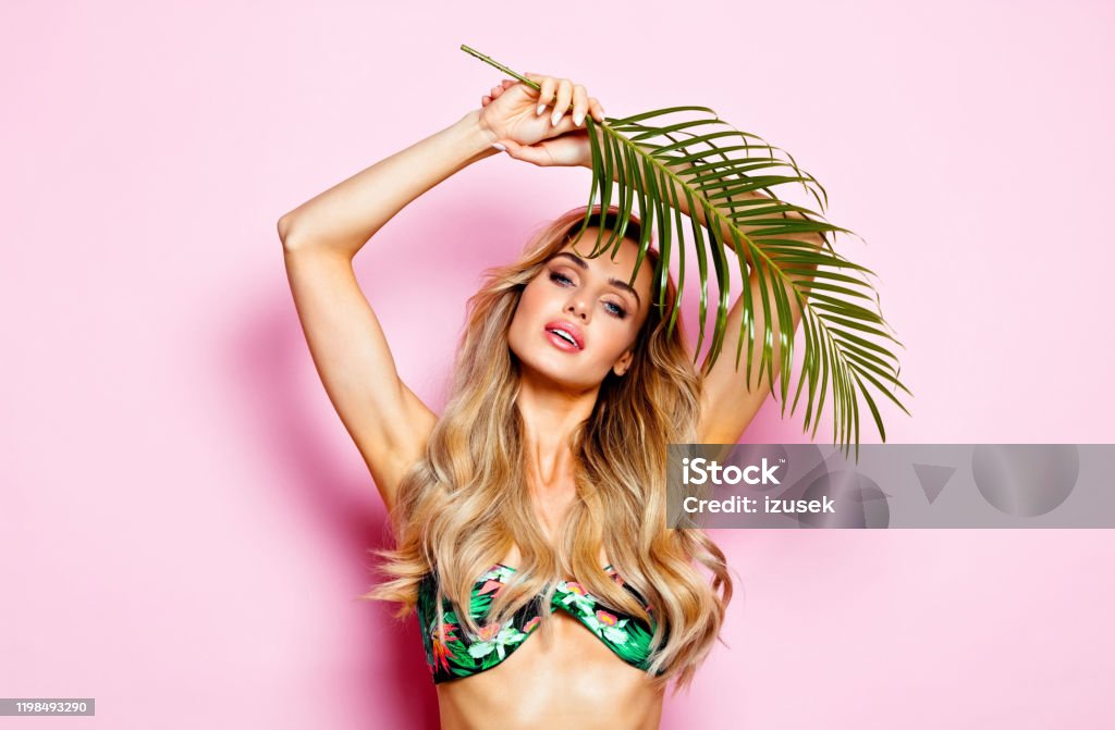 Summer portrait of young blonde woman in swimsuit Glamour portrait of beautiful long hair young woman wearing floral swimsuit, holding palm leaves. Studio shot on pink background. 25-29 Years Stock Photo