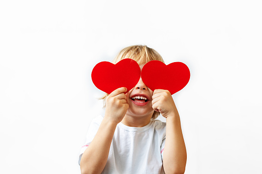 A little boy in a white shirt holds red hearts in his hands, closes his eyes and smiles on a white background. Valentines Day, Mothers Day, International Womens day