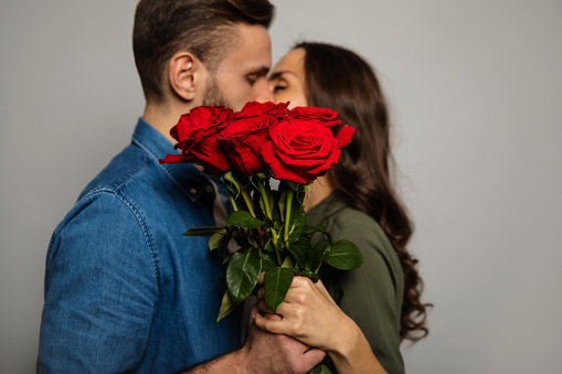Close-up photo of a lovely couple in casual outfits, who are hiding behind a bouquet of red roses while kissing.