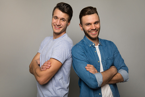 The strength of brothers. Two young men in casual clothes are standing back to back with cheerful facial expressions, posing with folded arms and smiling to the camera.