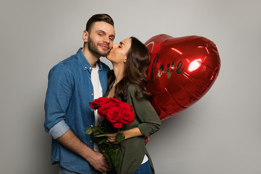 Сlose-up photo of a charming lady, who is kissing her attractive boyfriend in the cheek, while holding a bouquet of roses and a big red heart-shaped balloon.