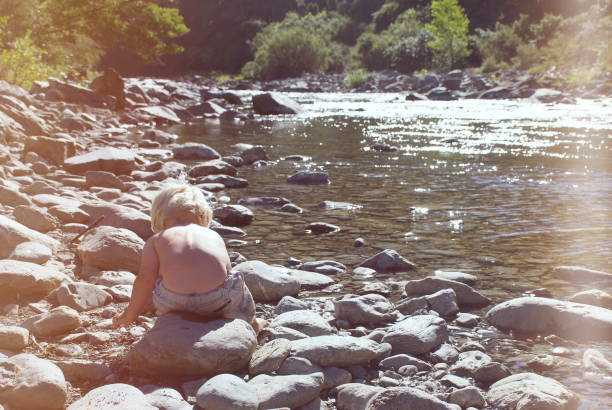 A young toddler boy plays with rocks by the river in summer.