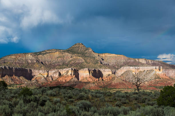 Photo of Storm clouds and faint rainbows over a desert mountain peak with colorful cliffs highlighted by the light of sunset