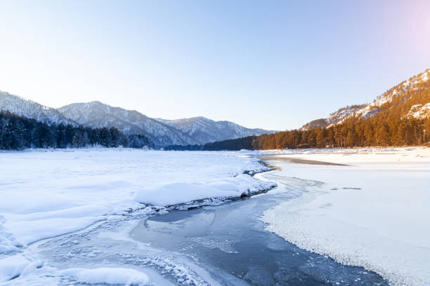 paisaje invernal con un río katun congelado o un lago en las montañas altai en un día soleado bajo un cielo azul con nieve a la deriva en un círculo dividido en lados cálidos y fríos por un arroyo. - siberia river nature photograph fotografías e imágenes de stock