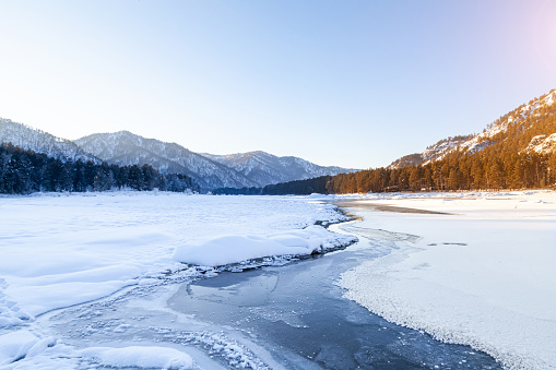 Winter landscape with a frozen Katun river or a lake in the Altai mountains on a sunny day under a blue sky with snowdrifts in a circle divided into warm and cold sides by a stream.